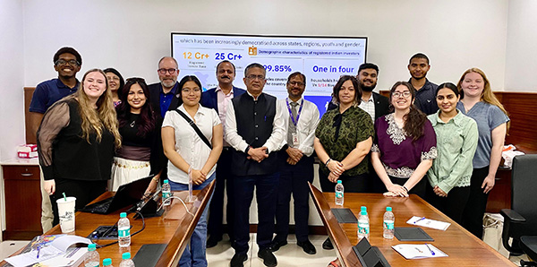 A group photo of MGA students and faculty standing with Shri Ashishkumar Chauhan and other officials in a conference room at the National Stock Exchange of India. A digital presentation about Indian investor demographics is visible on the screen in the background.